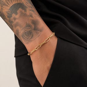 May include: A gold-toned chain bracelet featuring a series of elongated oval links alternating with smaller, textured cylindrical segments. The bracelet has a polished finish and a secure clasp mechanism, worn around the wrist.