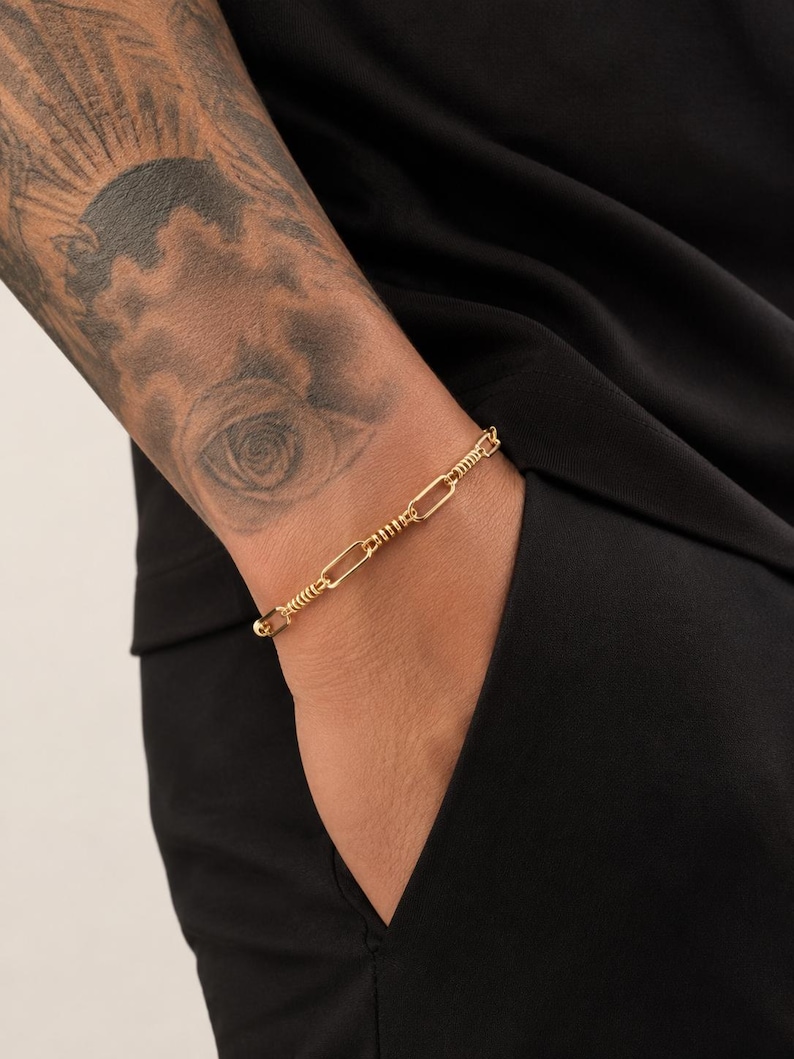 May include: A gold-toned chain bracelet featuring a series of elongated oval links alternating with smaller, textured cylindrical segments. The bracelet has a polished finish and a secure clasp mechanism, worn around the wrist.