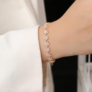 May include: A gold-toned bracelet featuring a floral design. Each flower is adorned with small, clear crystals. The bracelet is worn on a wrist, with a white sleeve in the background.