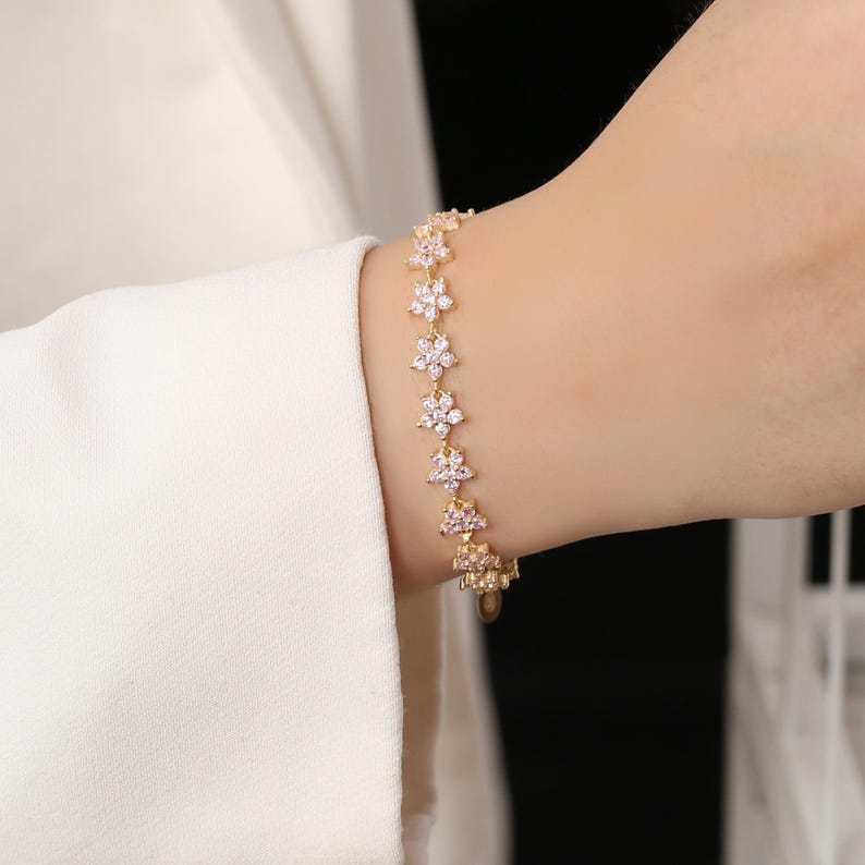 May include: A gold-toned bracelet featuring a floral design. Each flower is adorned with small, clear crystals. The bracelet is worn on a wrist, with a white sleeve in the background.