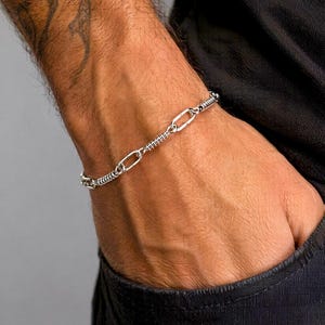 May include: A silver-toned chain bracelet featuring a repeating pattern of elongated oval links alternating with sections of tightly coiled wire. The metallic links have a polished finish and are connected in a continuous, flexible loop design.