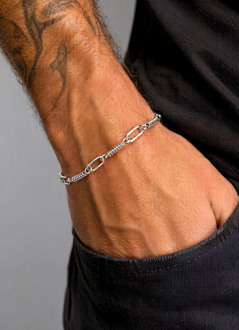 May include: A silver-toned chain bracelet featuring a repeating pattern of elongated oval links alternating with sections of tightly coiled wire. The metallic links have a polished finish and are connected in a continuous, flexible loop design.