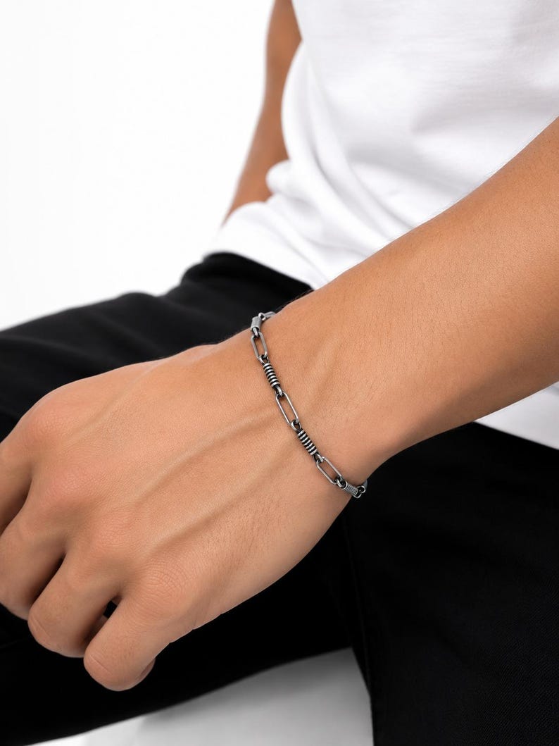 May include: A silver-toned metal chain bracelet featuring alternating elongated oval links and smaller, textured cylindrical segments with a dark, oxidized finish, creating a distinct industrial appearance on the wrist.