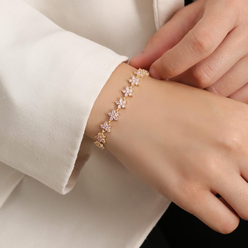 May include: A gold-colored bracelet featuring a series of flower-shaped links. Each flower has small, pink-colored accents. The bracelet is worn on a wrist, and the clasp is not visible. The background is a white shirt.