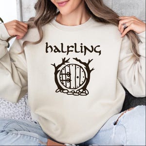 May include: A cream colored sweatshirt with the word "halfling" printed in black cursive lettering. Below the word is a black and white illustration of a round door with a wooden frame and a small window. The door is surrounded by two trees and rocks.