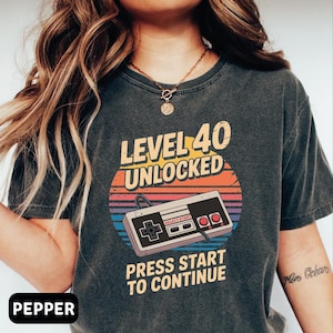 May include: Dark gray t-shirt with a retro video game controller graphic. The design includes the text "LEVEL 40 UNLOCKED" above the controller and "PRESS START TO CONTINUE" below. The graphic features a sunset-style background with orange, yellow, blue, and purple stripes.