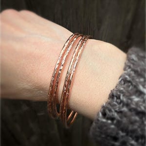 May include: Four copper-toned bangles with a textured, hammered finish. The bracelets are worn on a wrist, displaying their warm metallic colour. The background is blurred, highlighting the jewellery.