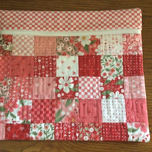 May include: A quilted patchwork zippered pouch in shades of red and pink, featuring floral, heart, and checkered patterns. The top edge has a red and white checkered design. A decorative flower charm is attached to the zipper.