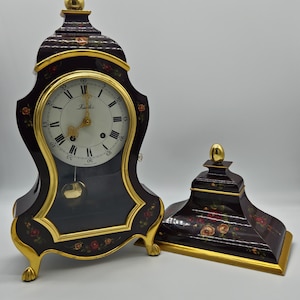 May include: A dark brown mantel clock with gold trim and floral accents, featuring a white face with Roman numerals and a gold pendulum. Beside it sits a matching dark brown rectangular base with gold trim and painted floral details.