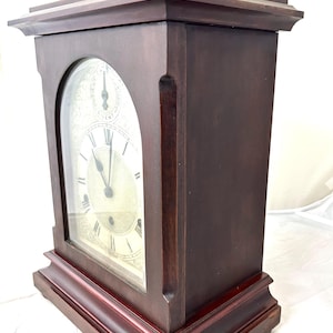 Puede incluir: Un reloj de chimenea de madera marrón oscuro con cuerpo rectangular y esfera de cristal arqueada. La esfera del reloj muestra números romanos y dos esferas más pequeñas. Un elemento decorativo dorado se encuentra en la parte superior.