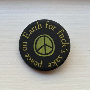 May include: Black button with a peace sign and the text "Peace on Earth for fuck's sake" in yellow.