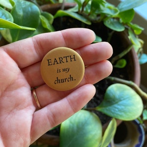 K&ouml;nnte beinhalten: Ein goldener Knopf mit dem Text "EARTH is my church." Der Knopf wird von einer Hand gehalten.