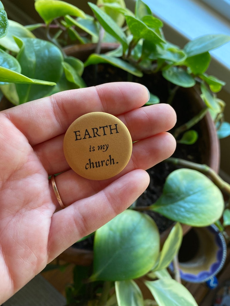 K&ouml;nnte beinhalten: Ein goldener Knopf mit dem Text "EARTH is my church." Der Knopf wird von einer Hand gehalten.
