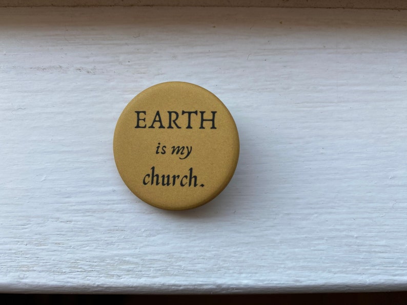 K&ouml;nnte beinhalten: Ein runder, goldfarbener Knopf mit dem Text "EARTH is my church." in schwarzer Schrift.