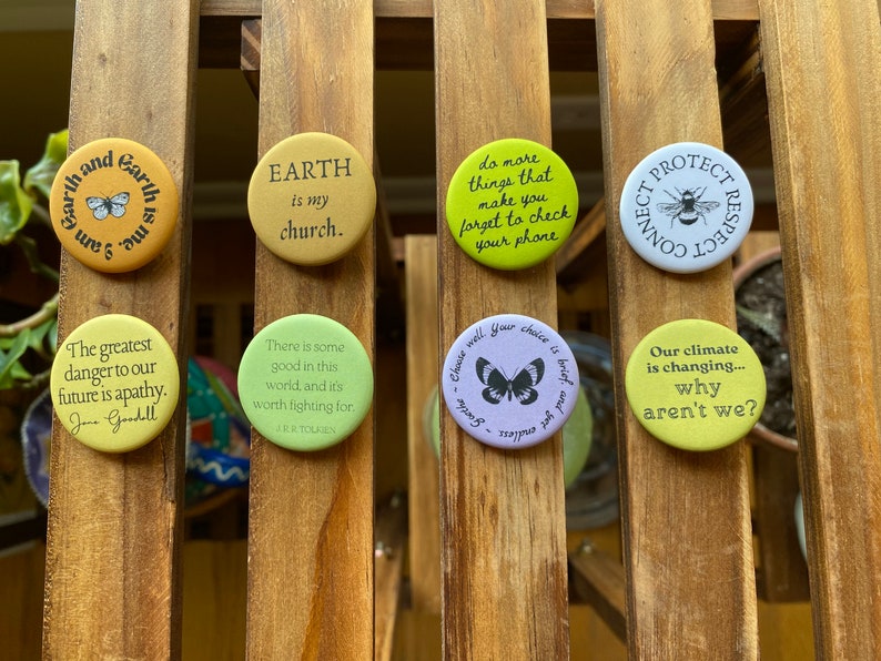 K&ouml;nnte beinhalten: Eine Sammlung von neun runden Anstecknadeln mit verschiedenen Umwelt- und inspirierenden Botschaften. Die Anstecknadeln zeigen einen Schmetterling, eine Biene und Texte wie "Die Erde ist meine Kirche", "Unser Klima ver&auml;ndert sich... warum nicht wir?" und "Die gr&ouml;&szlig;te Gefahr f&uuml;r unsere Zukunft ist Apathie."
