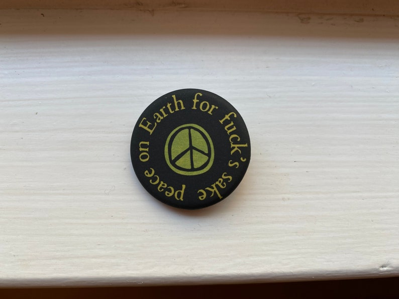 K&ouml;nnte beinhalten: Schwarzer Knopf mit einem Peace-Zeichen und dem Text "Peace on Earth for fuck's sake" in Gelb.