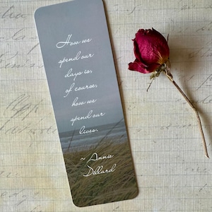 May include: A rectangular bookmark with rounded corners, featuring a quote and author's name. The bookmark has a gray gradient background with a coastal scene at the bottom. A dried red rose rests beside the bookmark on a textured surface.
