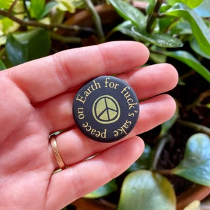 K&ouml;nnte beinhalten: Schwarzer Knopf mit einem Peace-Zeichen und dem Text "Peace on Earth for fuck's sake" in Gelb.