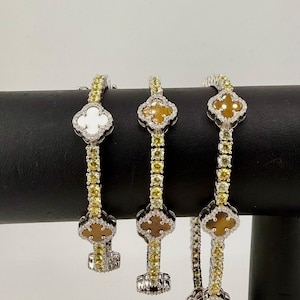 May include: Three elegant bracelets with clover-shaped pendants. The bracelets feature yellow and white gemstones, with a mix of yellow and white clover designs. The bracelets are displayed on a black stand against a white background.