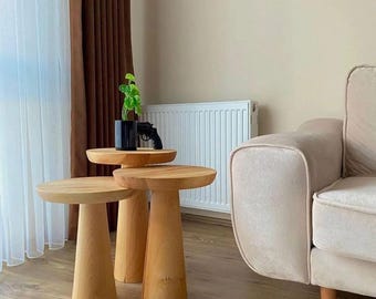 May include: Three wooden nesting tables with a natural finish. The tables are round and have a wide, flared base. The tables are arranged in a staggered formation, with the largest table in the back and the smallest table in the front.