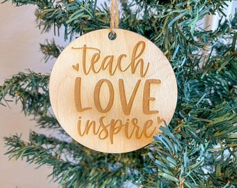 May include: A round wooden ornament with the text "teach love inspire" engraved in a light brown color. The ornament is hanging from a string on a green artificial Christmas tree.