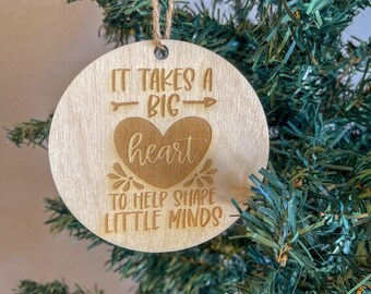 May include: A round wooden ornament with the text "It takes a big heart to help shape little minds". The ornament is decorated with a heart and an arrow.