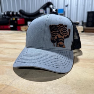 Op de afbeelding: Een grijze en zwarte truckercap met een bruine leren patch met een cartoonbeer die een Amerikaanse vlag vasthoudt.