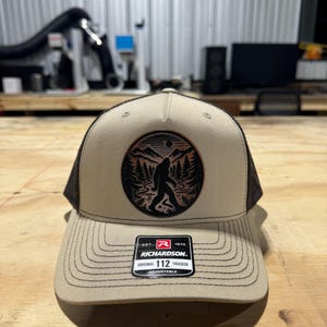 Op de afbeelding: Een beige en zwarte trucker-pet met een bruine leren patch met een silhouet van Bigfoot die door een bos met bergen op de achtergrond loopt. De patch heeft de tekst "EST. 1970 RICHARDSON. ORIGINAL 112 TRUCKER ADJUSTABLE".