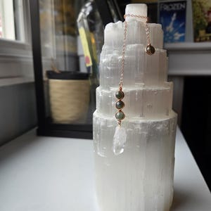 May include: A white selenite tower with a rose gold chain necklace featuring green beads and a clear crystal pendant. The necklace is draped over the tower. The background includes a window and decorative items. The tower is approximately 15 cm tall.