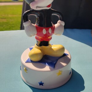 May include: A ceramic Mickey Mouse music box with a white base decorated with blue and yellow stars. Mickey is standing on a blue star with his hands on his hips. He is wearing his signature red shorts with two black buttons and yellow shoes.