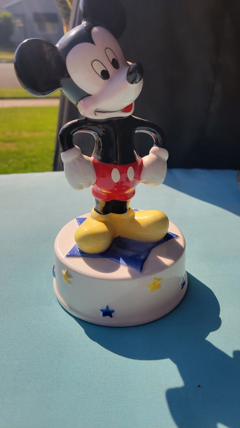 May include: A ceramic Mickey Mouse music box with a white base decorated with blue and yellow stars. Mickey is standing on a blue star with his hands on his hips. He is wearing his signature red shorts with two black buttons and yellow shoes.