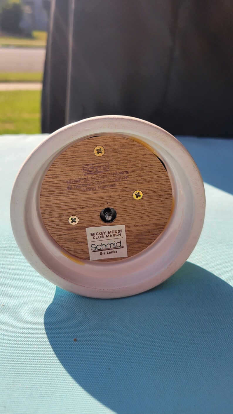May include: A white ceramic music box with a wooden base. The base has a label that reads "Mickey Mouse Club March Schmid Sri Lanka". The music box is hand-painted and features a Mickey Mouse design.