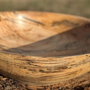 May include: A handcrafted wooden bowl with a natural, organic aesthetic. The bowl is made from light-colored wood with dark grain patterns, showcasing the wood's natural beauty. The bowl is approximately 10 inches in diameter and 3 inches deep.
