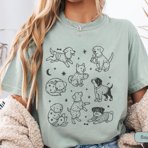 May include: Sage green t-shirt featuring a collection of illustrated dogs in various poses. The black and white drawings include dogs with bones, coffee cups, and star and moon accents. The shirt has a relaxed fit and short sleeves.
