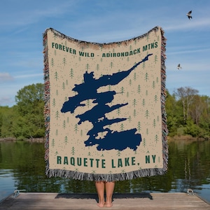 May include: A beige throw blanket with a dark blue lake design and green text that reads "FOREVER WILD - ADIRONDACK MTNS" and "RAQUETTE LAKE, NY." The blanket has a fringed edge and a pattern of small green trees.