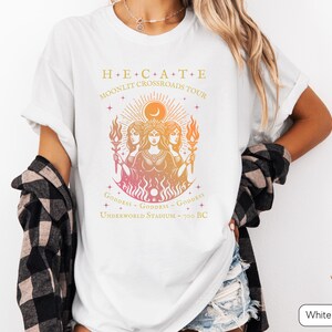A vintage-style Hecate goddess T-shirt featuring a triple goddess illustration, inspired by Greek mythology.  The  text says “Hecate Moonlit Crossroads Tour” and “Goddess - Goddess - Goddess” with a gradient of warm colors.