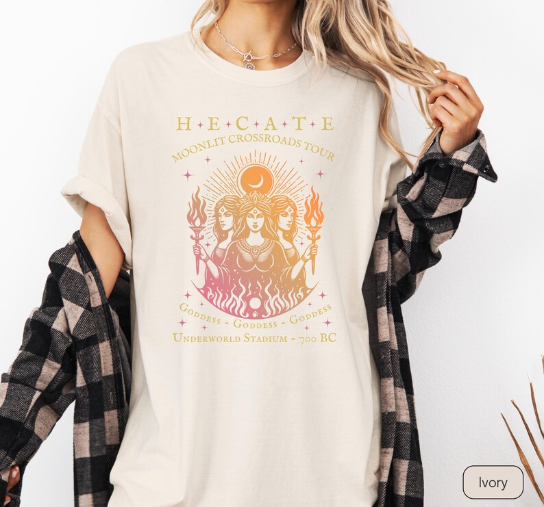 A vintage-style Hecate goddess T-shirt featuring a triple goddess illustration, inspired by Greek mythology.  The  text says “Hecate Moonlit Crossroads Tour” and “Goddess - Goddess - Goddess” with a gradient of warm colors.