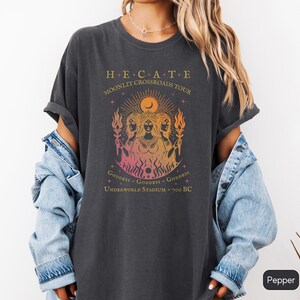 A vintage-style Hecate goddess T-shirt featuring a triple goddess illustration, inspired by Greek mythology.  The  text says “Hecate Moonlit Crossroads Tour” and “Goddess - Goddess - Goddess” with a gradient of warm colors.