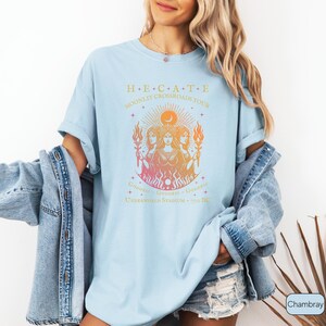 A vintage-style Hecate goddess T-shirt featuring a triple goddess illustration, inspired by Greek mythology.  The  text says “Hecate Moonlit Crossroads Tour” and “Goddess - Goddess - Goddess” with a gradient of warm colors.