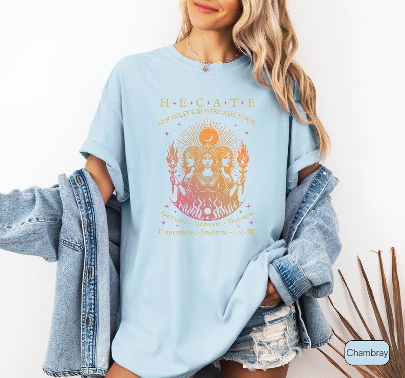 A vintage-style Hecate goddess T-shirt featuring a triple goddess illustration, inspired by Greek mythology.  The  text says “Hecate Moonlit Crossroads Tour” and “Goddess - Goddess - Goddess” with a gradient of warm colors.