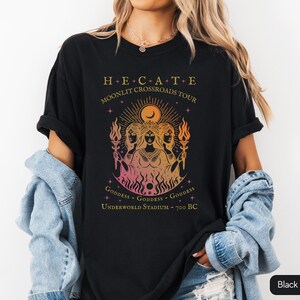 A vintage-style Hecate goddess T-shirt featuring a triple goddess illustration, inspired by Greek mythology.  The  text says “Hecate Moonlit Crossroads Tour” and “Goddess - Goddess - Goddess” with a gradient of warm colors.