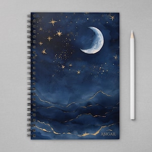 May include: A blue spiral notebook with a watercolor design of a crescent moon and stars against a dark blue background. The notebook has gold accents and the word "ABIGAIL" printed on the bottom right corner.