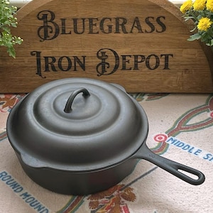 Op de afbeelding: Een zwarte gietijzeren koekenpan met deksel en handvat, met de tekst "Bluegrass Iron Depot" op een houten bord. De koekenpan staat op een oppervlak met patroon en tekst.