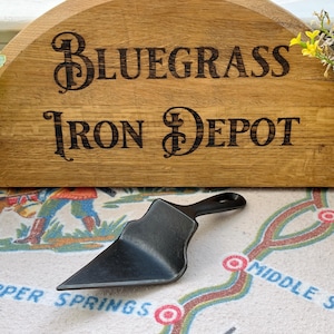 May include: A black cast iron serving spatula with a triangular blade and a short handle. The spatula is positioned in front of a curved wooden sign featuring the burned-in text "Bluegrass Iron Depot" in a decorative serif font style.