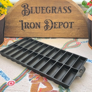 Puede incluir: Molde negro de hierro fundido para pan de maíz con moldes rectangulares. La sartén está sobre un paño estampado, con un letrero de madera en el fondo que dice "Bluegrass Iron Depot". Dos cestas de flores flanquean el letrero.