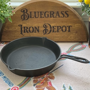 Op de afbeelding: Een zwarte gietijzeren koekenpan met een handvat, gepresenteerd voor een houten bord met de tekst "Bluegrass Iron Depot". De pan staat op een patroon oppervlak, met potplanten aan weerszijden.