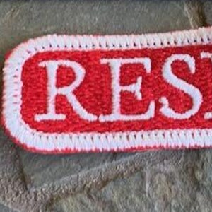 以下が含まれることがあります： 白いステッチと白い文字で「RESIST」と書かれた赤い布のパッチ