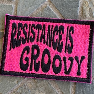 Può includere: Un cerotto in tessuto rosa con testo ricamato nero che recita "Resistance is groovy".