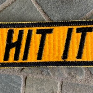 May include: A yellow and black embroidered patch with the text "HIT IT" in black.
