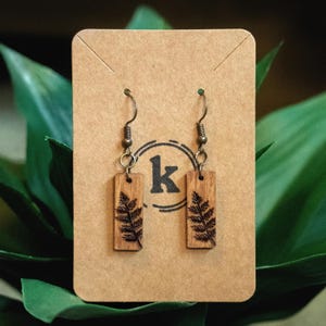 May include: Wooden dangle earrings with a fern design etched into rectangular wooden pendants. The earrings hang from bronze-colored hooks and are displayed on a brown card with a circular logo.